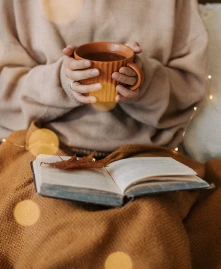 person holding mug with blanket and book on lap